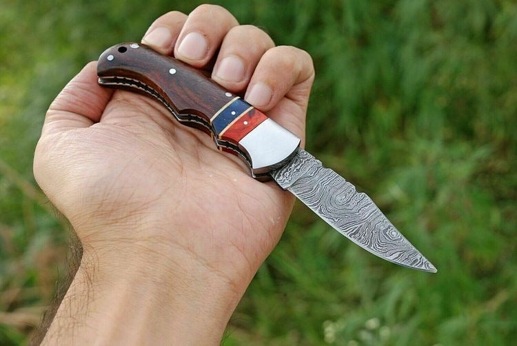 Artisanal Damascus folding knife with a 3-inch blade, ergonomic rosewood handle, and sturdy steel bolster, displayed with its leather sheath