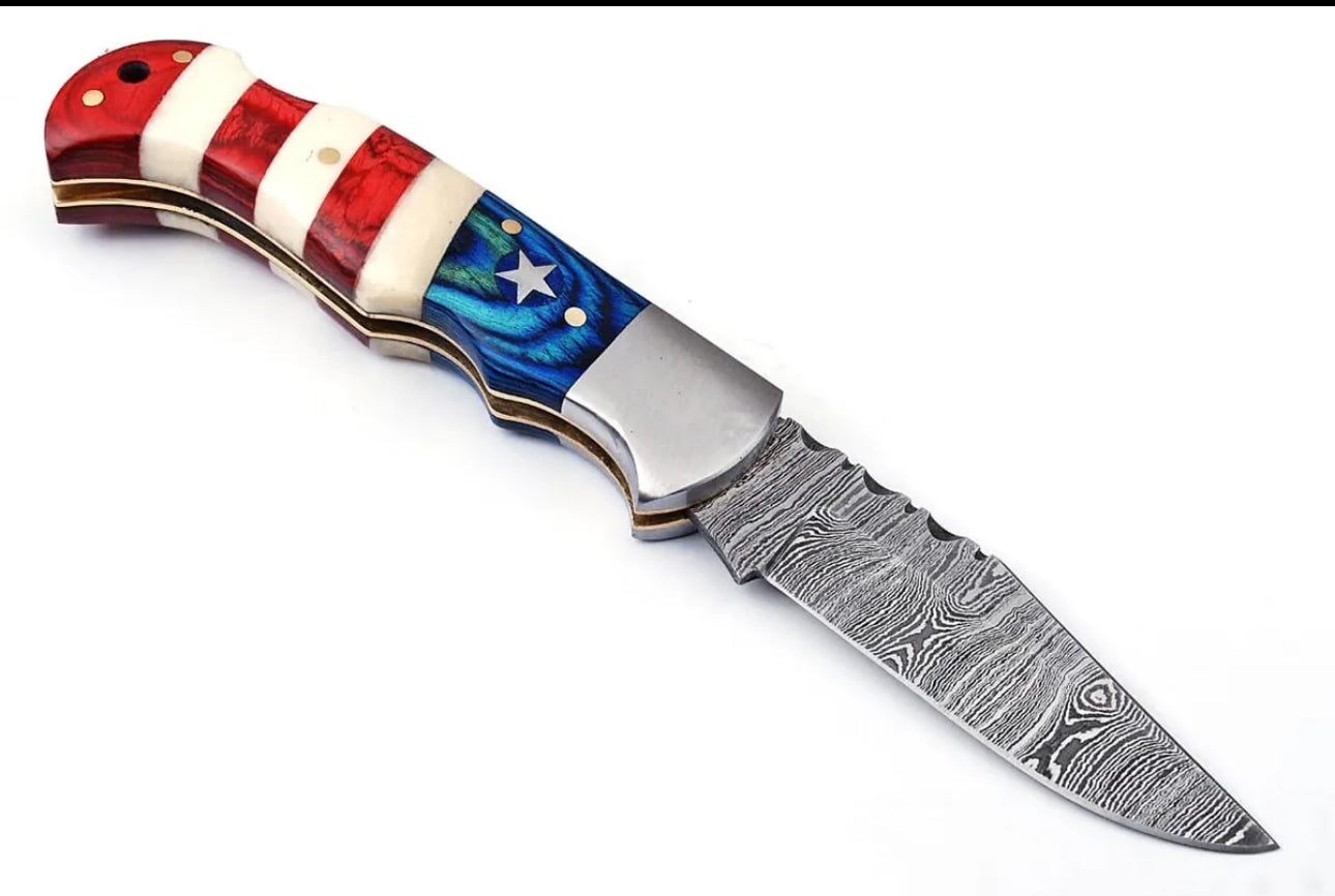 Close-up of a 3-inch Damascus steel folding knife with an American Flag handle and steel bolster, alongside a leather sheath.