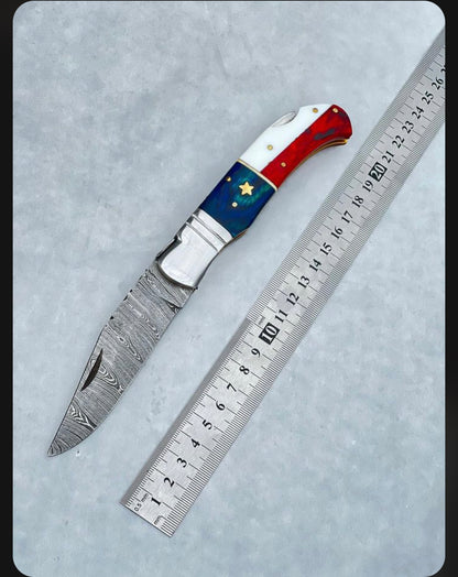 Compact Damascus folding knife with a 3-inch blade, American flag handle, steel bolster, and leather sheath for easy carry